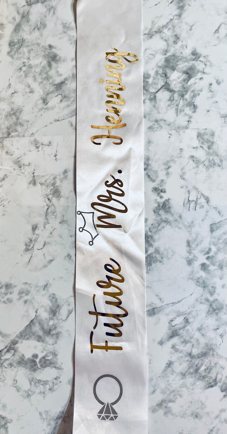 Puede incluir: Banda de sat&eacute;n blanco con letras doradas que dicen "Future Mrs. [Last Name]" con una corona y un gr&aacute;fico de anillo de diamantes.
