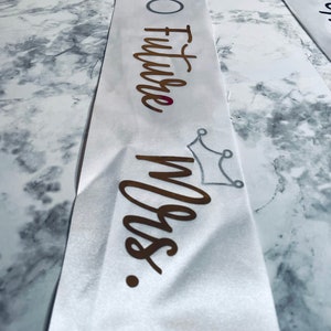 Puede incluir: Banda de sat&eacute;n blanco con letras doradas que dicen "Future Mrs." y un gr&aacute;fico de anillo de diamantes. Un gr&aacute;fico de corona plateada est&aacute; debajo del texto.