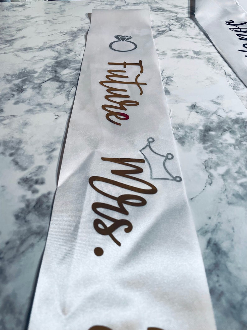 Puede incluir: Banda de sat&eacute;n blanco con letras doradas que dicen "Future Mrs." y un gr&aacute;fico de anillo de diamantes. Un gr&aacute;fico de corona plateada est&aacute; debajo del texto.