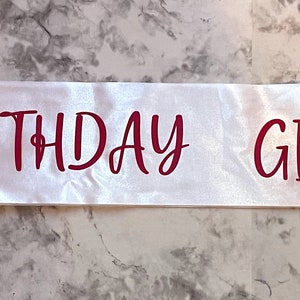 Puede incluir: Banda de sat&eacute;n blanco con las palabras "Birthday Girl" impresas en rojo.