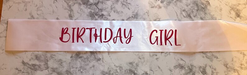 Puede incluir: Banda de sat&eacute;n blanco con las palabras "Birthday Girl" impresas en rojo.