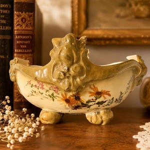 May include: A cream-coloured ceramic bowl with a scalloped, olive-green rim featuring raised scrollwork and three feet. The exterior is decorated with hand-painted orange sunflowers, small pink blossoms, and blue floral sprigs across the front.