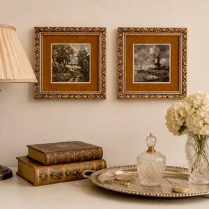 May include: A table lamp with a pleated beige shade, two square framed landscape prints with gold ornate borders, two stacked leather-bound books, a silver serving tray, a clear glass perfume bottle, and a clear glass vase with white flowers.