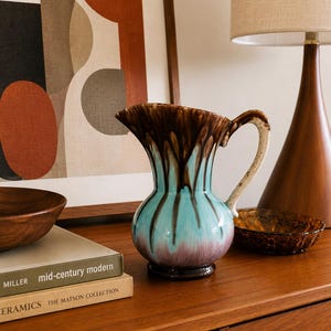 May include: A ceramic pitcher featuring a glossy glaze with a drip pattern in shades of dark brown, turquoise, and muted purple. The vessel has a curved handle, a flared rim, and a rounded body that tapers towards a circular base.