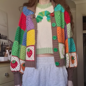 Puede incluir: Un cárdigan de ganchillo patchwork colorido con ribete marrón y botones verdes. Las mangas presentan cuadrados con fresas y naranjas. Debajo se lleva un suéter color crema con un diseño de oruga.