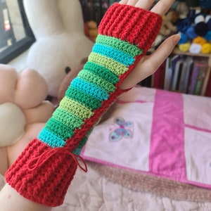 May include: A red, green, and blue striped crocheted fingerless glove with a red cuff. The glove is on a person's hand.