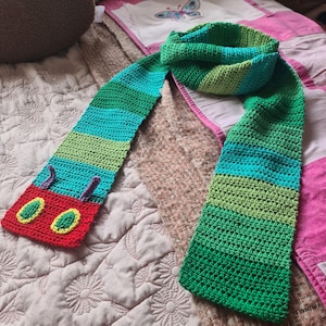 May include: A green, blue, and red striped crocheted scarf with a caterpillar face on one end. The caterpillar face has two large yellow eyes with green pupils and a red mouth.