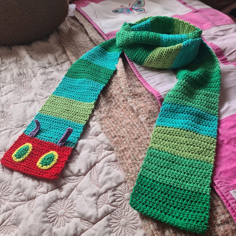 May include: A green, blue, and red striped crocheted scarf with a caterpillar face on one end. The caterpillar face has two large yellow eyes with green pupils and a red mouth.
