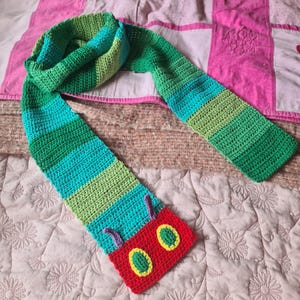 May include: A green, blue, and red crocheted scarf with a caterpillar design. The caterpillar has a red body with green and yellow eyes.