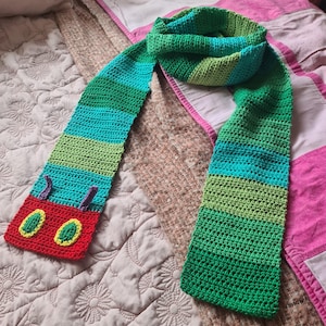 May include: A green, blue, and red striped crocheted scarf with a caterpillar face on one end. The scarf is made with a chunky yarn and has a simple stitch pattern.