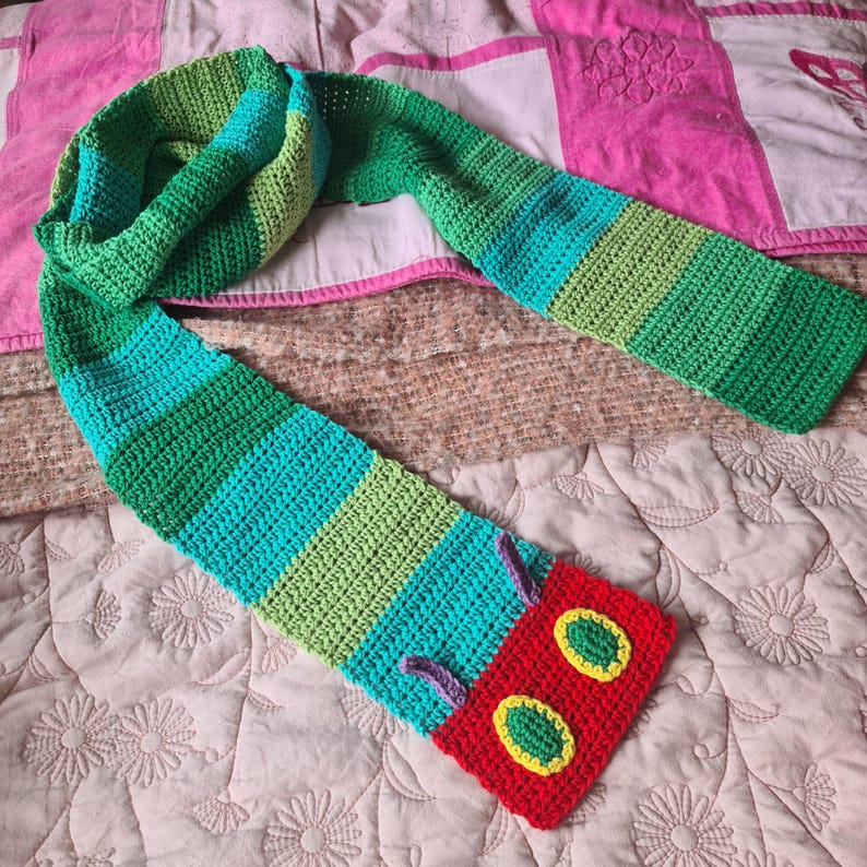 May include: A green, blue, and red crocheted scarf with a caterpillar face at the end. The caterpillar face has two large yellow eyes with green pupils and a red mouth.