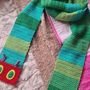 May include: A green, blue, and red crocheted scarf with a caterpillar applique. The caterpillar is red with green and yellow details.