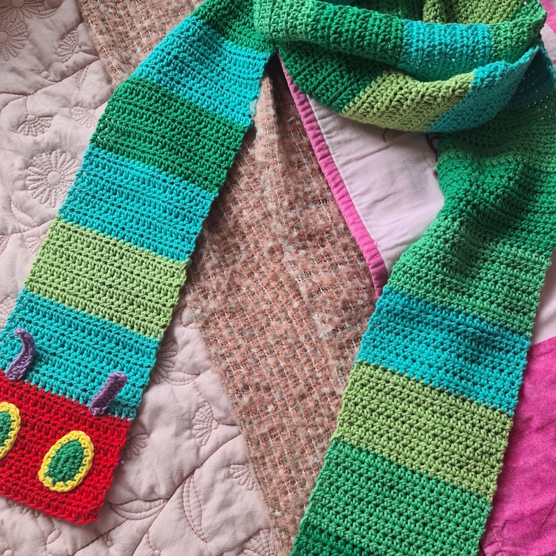 May include: A green, blue, and red crocheted scarf with a caterpillar applique. The caterpillar is red with green and yellow details.