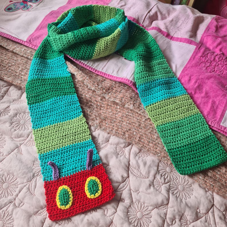 May include: A green, blue, and red crocheted scarf with a caterpillar design. The scarf has a red head with two yellow eyes and two purple antennae.