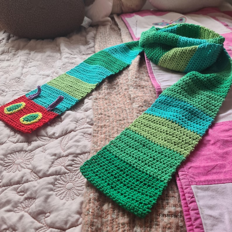 May include: A green, blue, and red crocheted scarf with a caterpillar face on one end. The scarf is made with a variety of stitches and is a fun and colorful accessory.