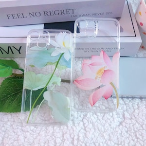 May include: Two clear phone cases with floral designs. The case on the left has a white lotus flower with green leaves. The case on the right has a pink lotus flower with green leaves. The text "FEEL NO REGRET" and "FAR AWAY, NEVER SEE." is visible on the box behind the cases.