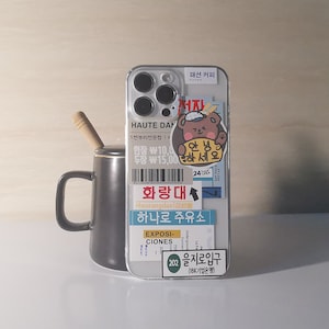 May include: A white iPhone case with various stickers, including a brown bear with a sailor hat and the text "안녕하세요" (Korean for "Hello"). The case also features a sticker with the text "화랑대" (Korean for "Hwarangdae") and "하나로 주유소" (Korean for "Hanaro Gas Station").