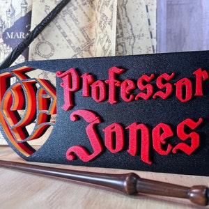 Peut inclure: Panneau noir avec le nom "Professor Jones" en lettres rouges de style gothique. Le panneau pr&eacute;sente un motif de bouclier superpos&eacute; avec des accents orange et jaune. Une baguette et d'autres articles th&eacute;matiques sont en arri&egrave;re-plan.