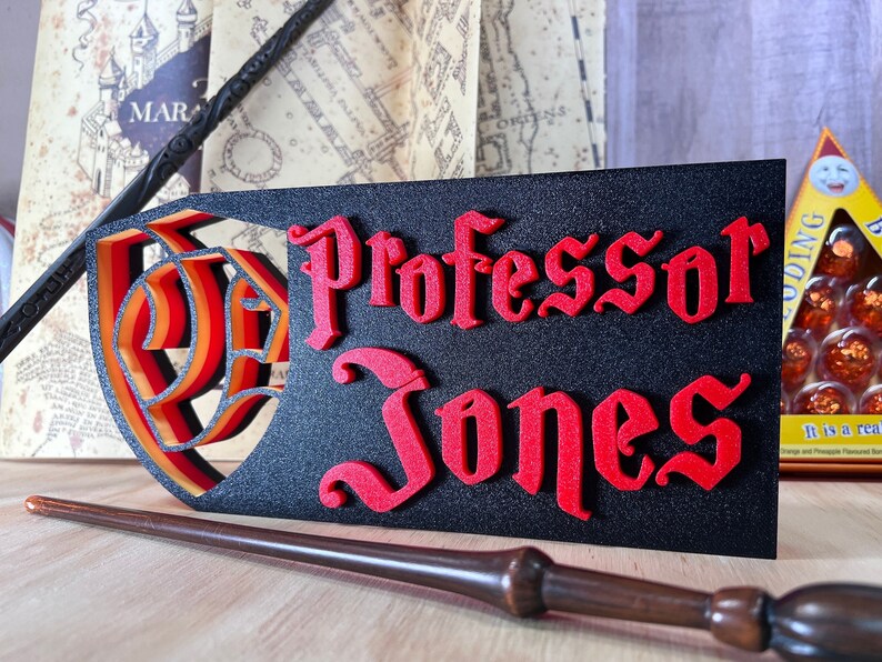 Peut inclure: Panneau noir avec le nom "Professor Jones" en lettres rouges de style gothique. Le panneau pr&eacute;sente un motif de bouclier superpos&eacute; avec des accents orange et jaune. Une baguette et d'autres articles th&eacute;matiques sont en arri&egrave;re-plan.