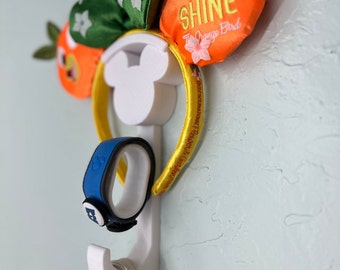 Peut inclure: Un crochet mural blanc en forme de Mickey Mouse avec un bandeau jaune avec le texte "Disneyland & California Adventure" et une paire d'oreilles de Minnie Mouse orange et vertes avec le texte "Hello Sunshine The Orange Bird" accrochés dessus. Un MagicBand bleu et un sac à main blanc sont également accrochés au crochet.