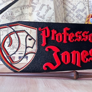 Peut inclure: Panneau rectangulaire noir avec le nom "Professor Jones" en rouge, lettrage stylis&eacute;. Un bouclier avec un embl&egrave;me de lion est &agrave; gauche. Une baguette repose en dessous, avec une carte en arri&egrave;re-plan.