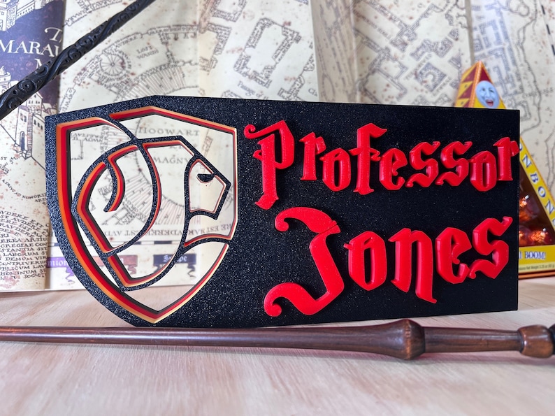 Peut inclure: Panneau rectangulaire noir avec le nom "Professor Jones" en rouge, lettrage stylis&eacute;. Un bouclier avec un embl&egrave;me de lion est &agrave; gauche. Une baguette repose en dessous, avec une carte en arri&egrave;re-plan.