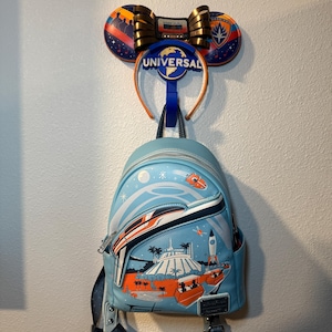 May include: A light blue backpack with a space-themed design, hanging below a pair of mouse ears and a headband with the word "UNIVERSAL". The backpack features a rocket, a monorail, and other space-themed elements.