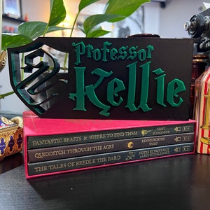Peut inclure: Un panneau noir avec le nom "Professor Kellie" en lettres vertes et un motif de bouclier vert. Le panneau est pos&eacute; sur une pile de livres et &agrave; c&ocirc;t&eacute; d'une bo&icirc;te de bonbons.