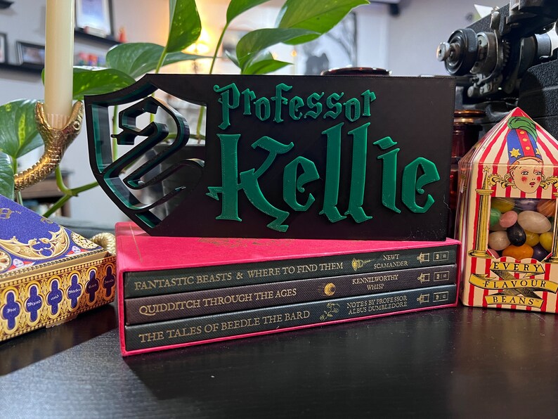 Peut inclure: Un panneau noir avec le nom "Professor Kellie" en lettres vertes et un motif de bouclier vert. Le panneau est pos&eacute; sur une pile de livres et &agrave; c&ocirc;t&eacute; d'une bo&icirc;te de bonbons.