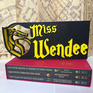 Peut inclure: Un panneau noir et jaune avec le nom "Miss Wendee" en lettres jaunes, orn&eacute; d'un embl&egrave;me stylis&eacute;. Le panneau repose sur une pile de livres rouges avec des titres tels que "Fantastic Beasts" et "Quidditch Through the Ages", &eacute;voquant un th&egrave;me fantastique.