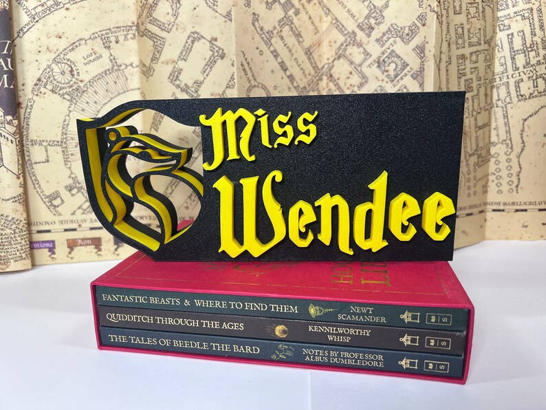 Peut inclure: Un panneau noir et jaune avec le nom "Miss Wendee" en lettres jaunes, orn&eacute; d'un embl&egrave;me stylis&eacute;. Le panneau repose sur une pile de livres rouges avec des titres tels que "Fantastic Beasts" et "Quidditch Through the Ages", &eacute;voquant un th&egrave;me fantastique.