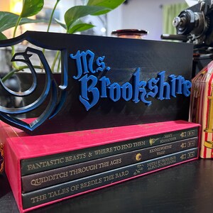 Peut inclure: Un panneau d&eacute;coratif noir et bleu avec le nom "Ms. Brookshire" en lettres bleues. Le panneau est plac&eacute; sur une pile de livres avec des titres tels que "Fantastic Beasts" et "Quidditch Through the Ages". Une bo&icirc;te de bonbons g&eacute;lifi&eacute;s est visible.