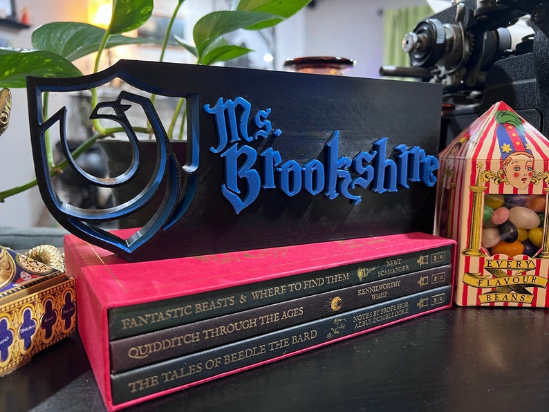 Peut inclure: Un panneau d&eacute;coratif noir et bleu avec le nom "Ms. Brookshire" en lettres bleues. Le panneau est plac&eacute; sur une pile de livres avec des titres tels que "Fantastic Beasts" et "Quidditch Through the Ages". Une bo&icirc;te de bonbons g&eacute;lifi&eacute;s est visible.