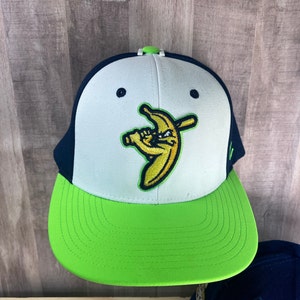 May include: A white and green baseball cap with a black brim. The front of the cap features an embroidered image of a banana holding a baseball bat. The banana is wearing a baseball cap and has a mischievous expression.