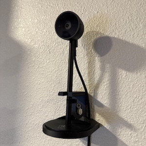 May include: A black webcam mounted on a wall. The webcam has a circular lens and is attached to a stand with a base and a cable. The stand is mounted on a textured, off-white wall.