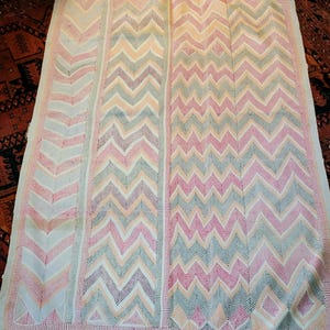 Op de afbeelding: Een groot, rechthoekig textiel met een herhalend zigzagpatroon in roze, blauwe en perzikkleuren op een crèmekleurige achtergrond. De randen zijn afgezet met een dun, rood gestreept patroon. Waarschijnlijk een deken of sprei.