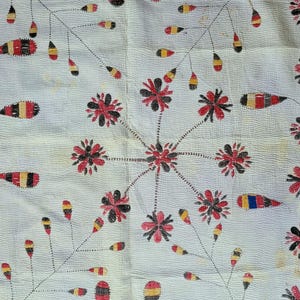 Op de afbeelding: Een crèmekleurig textiel met gedetailleerd handborduurwerk. De stof toont een herhalend patroon van zwarte en rode bloemmotieven verbonden door dunne lijnen, afgewisseld met gele, rode en zwarte druppelvormige elementen.