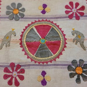 Puede incluir: Una pieza textil con un motivo circular central de segmentos alternos rosas y grises, rodeado por un borde festoneado rosa. La tela muestra flores bordadas grises y rosas, pequeñas flores moradas y amarillas, y dos figuras de animales grises.