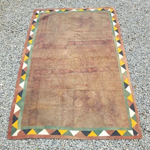 Op de afbeelding: Een grote, rechthoekige bruine quilt met een patroon. De quilt heeft een rand van afwisselende driehoeken in zwart, geel, wit en groen. De quilt is gemaakt van een zacht, getextureerd materiaal.