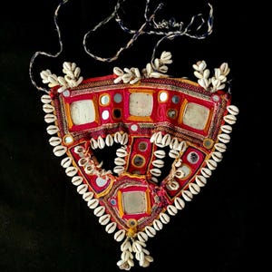 Puede incluir: Un collar de forma triangular con una base de tela roja y una variedad de adornos coloridos, incluyendo pequeños espejos, cuentas y conchas.