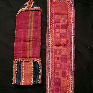 Op de afbeelding: Twee antieke textielstukken. Eén is een lange, smalle buidel met een flap, met rode en bruine strepen en blauwe en rode accenten. De andere is een rechthoekig paneel met geborduurde vierkanten in rood- en roze tinten.