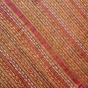 Könnte beinhalten: Nahaufnahme eines Textils mit einem sich wiederholenden Muster aus diagonalen Linien. Die Linien sind hauptsächlich braun, mit goldenen und weißen Akzenten. Ein tiefrosa Streifen verläuft diagonal über den Stoff.