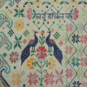 以下が含まれることがあります： クリーム色の生地に施されたクロスステッチ刺繍のクローズアップ。デザインは、ピンク、黄色、赤の花に囲まれた、向かい合う2羽の青い孔雀が特徴です。生地には外国語のテキストも含まれています。