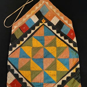 Puede incluir: Un tapiz de tela acolchada con un patrón central de diamantes compuesto por triángulos amarillos, azules, verdes y color melocotón. El borde muestra un diseño geométrico en zigzag negro, blanco y naranja, con un lazo de cordón blanco.