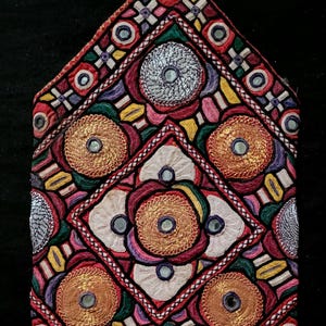 Peut inclure: Textile brodé aux couleurs vives avec un motif géométrique. Le design comprend des coutures complexes en rouge, or, blanc et violet, avec de petits accents miroir. La forme générale est un triangle stylisé, probablement un élément décoratif.
