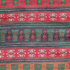 Könnte beinhalten: Ein gewebtes Textil mit horizontalen Streifen in Rot, Dunkellila und Orange. Das Muster besteht aus sich wiederholenden grünen geometrischen Formen, die an Säulen erinnern, mit kleinen roten Rautenakzenten, abwechselnd mit Reihen aus weißen und grünen Rautenmotiven.