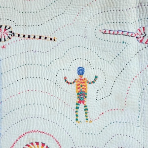 Op de afbeelding: Een wit textiel met ingewikkeld kantha-borduurwerk met een herhalend patroon van golvende lijnen. De stof toont kleurrijke geborduurde motieven, waaronder een centrale menselijke figuur, cirkelvormige vormen en rode, blauwe en gele lijnen.