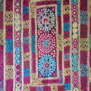 May include: A rectangular textile wall hanging featuring concentric borders of embroidered geometric patterns in yellow, blue, pink, and red. The design includes circular mirrors embedded in the borders and three central floral motifs.