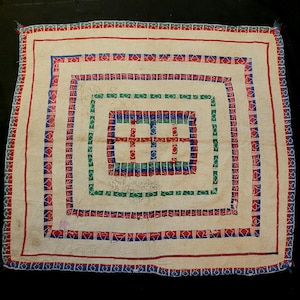 Op de afbeelding: Crèmekleurig textiel met een vierkant ontwerp. Het textiel heeft concentrische vierkante randen in rood, blauw en groen, met geometrische patronen. De buitenste rand is rood, gevolgd door blauwe en groene vierkanten. Het midden heeft een rood en groen kruis.