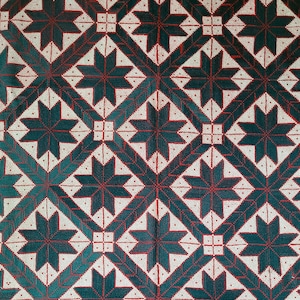Op de afbeelding: Een textielstof met een herhalend geometrisch patroon van donkergroene achtpuntige sterren in het midden van ruitvormen. Het ontwerp is omlijnd met dunne rode lijnen en kleine rode stippen op een crèmekleurige achtergrond, wat een raster vormt.